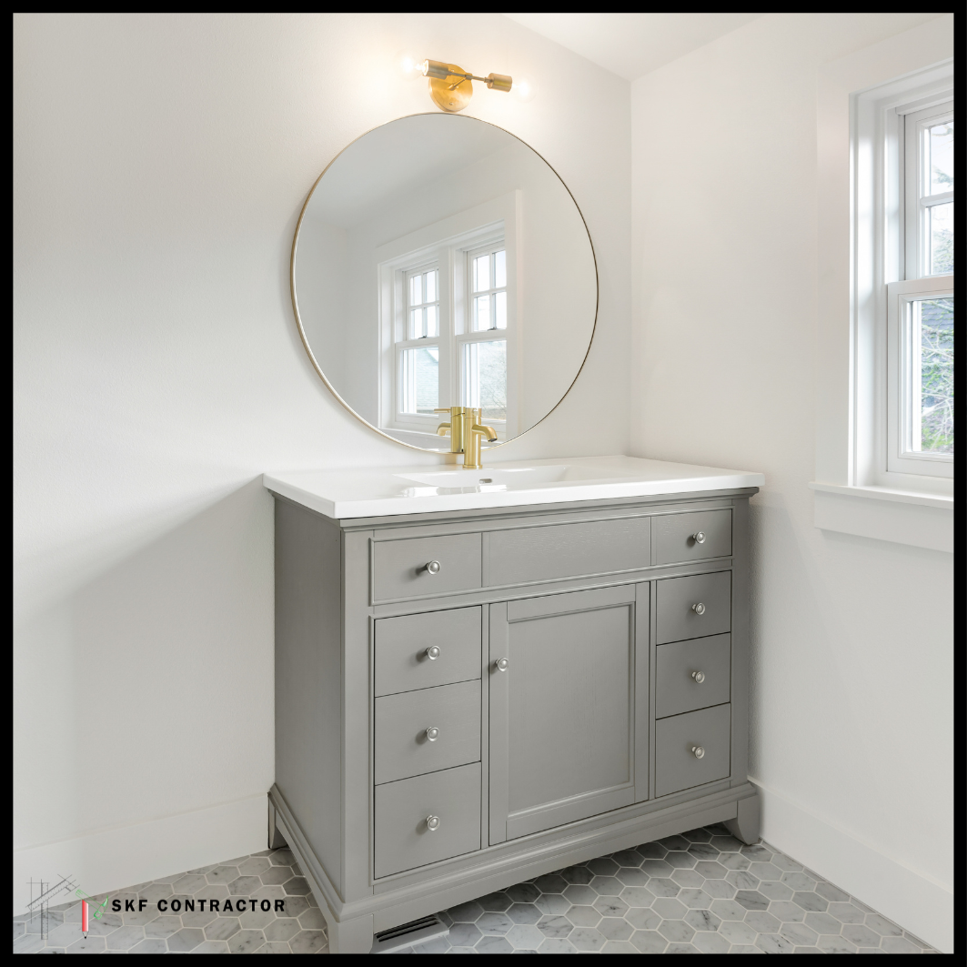 Grey bathroom vanity with white countertop, gold round mirror, brass sconce, and hexagon marble tile floor.