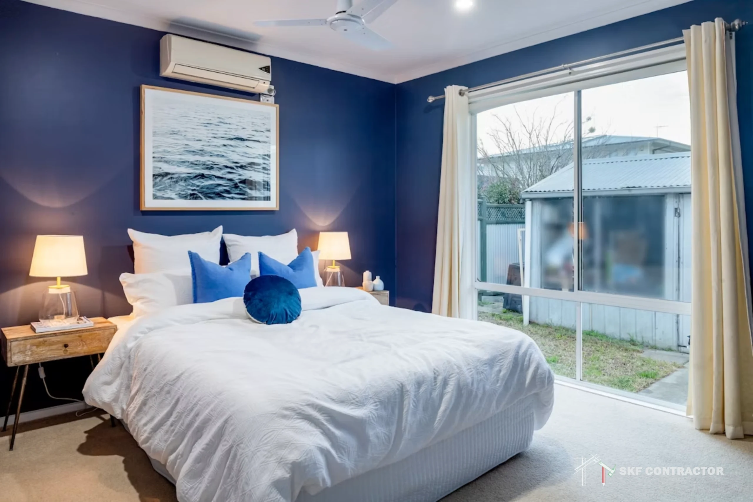 Bedroom with navy blue walls, white bedding, two bedside tables with lamps, a large window with cream curtains, and a framed picture above the bed.