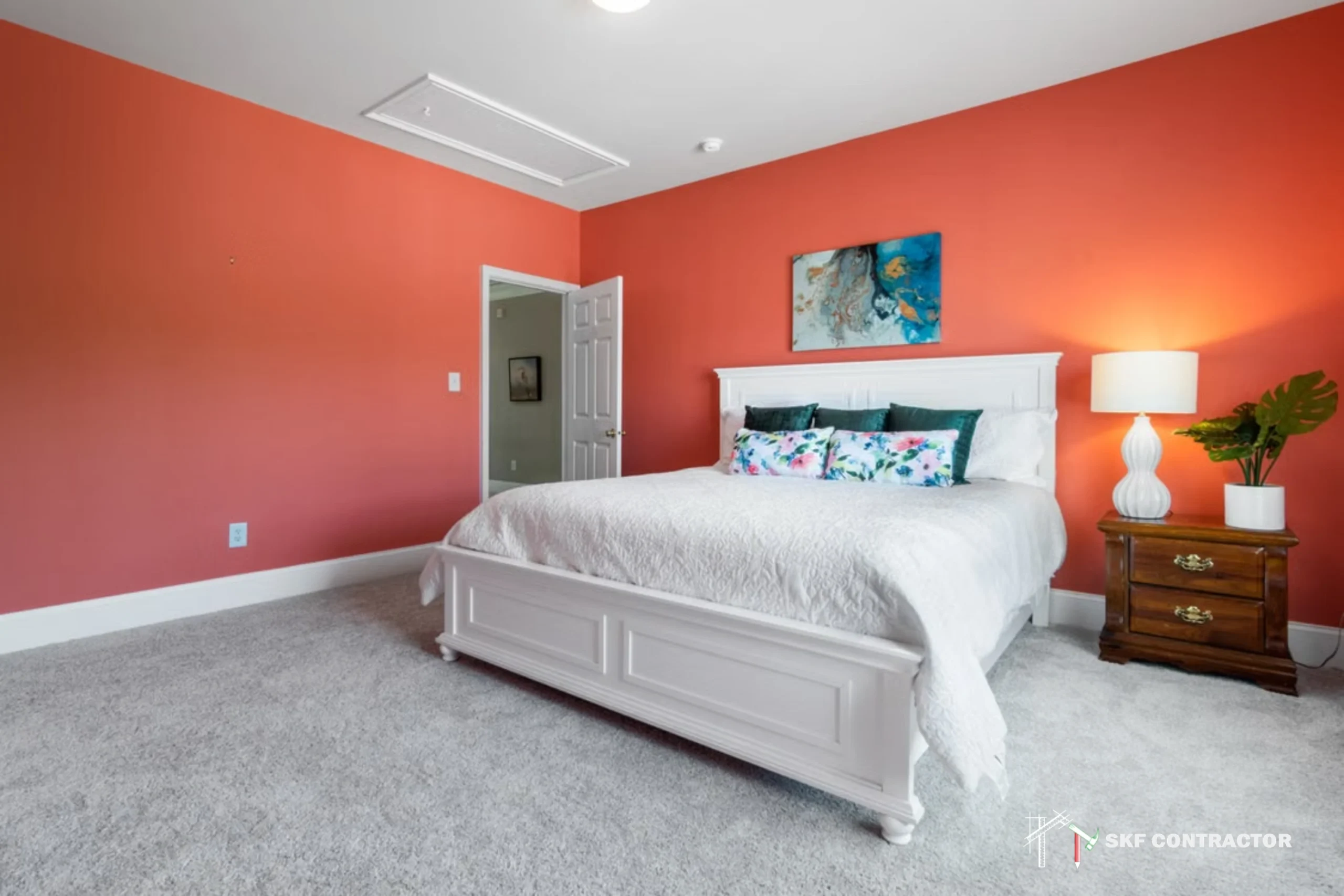 Bright coral bedroom with a white bed, floral pillows, gray carpet, artwork, wood nightstand, and glowing lamp; inviting and peaceful.