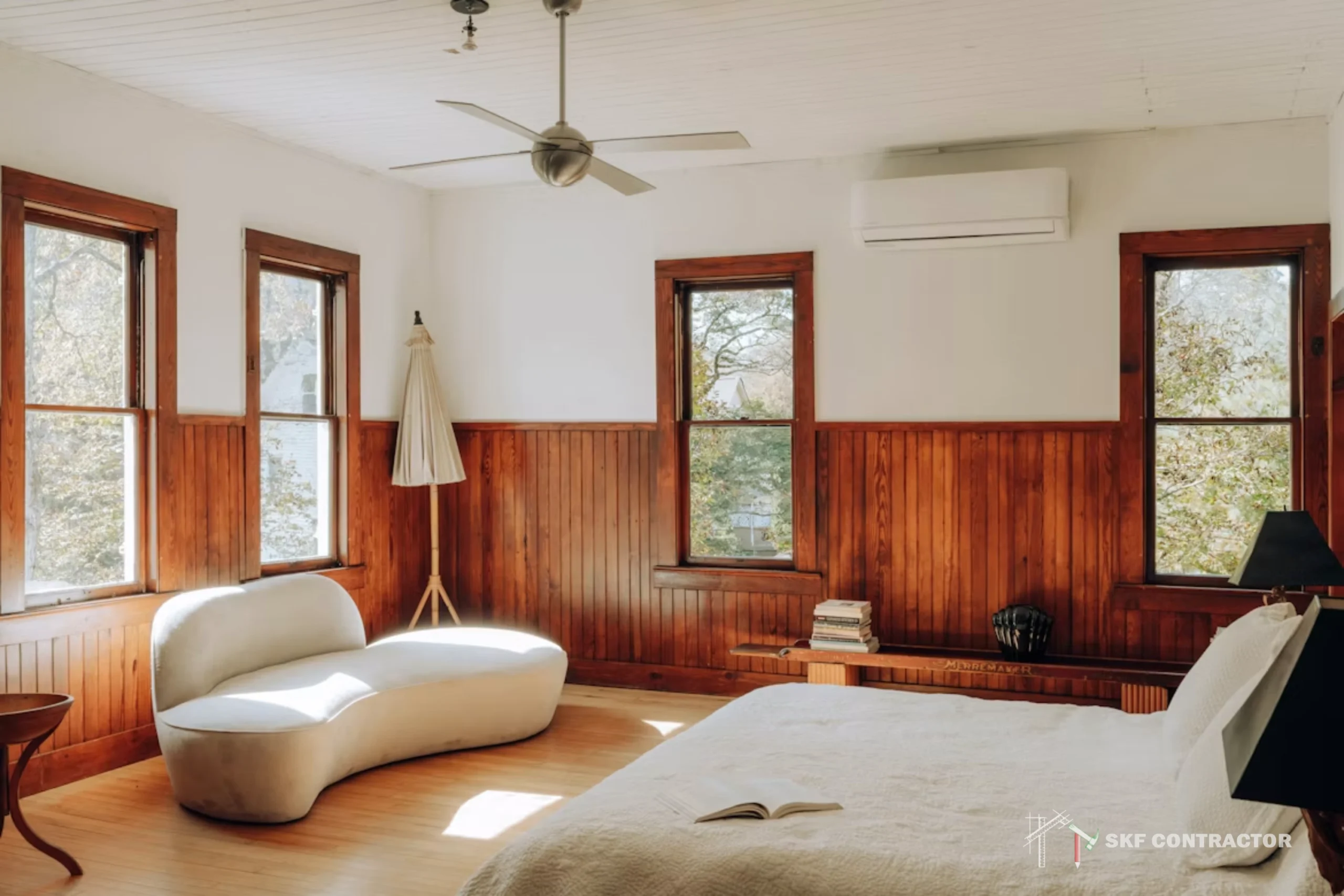 Sunlit bedroom with wood-paneled walls, tall windows, a curved white chair, neatly made bed, ceiling fan, and serene natural views.